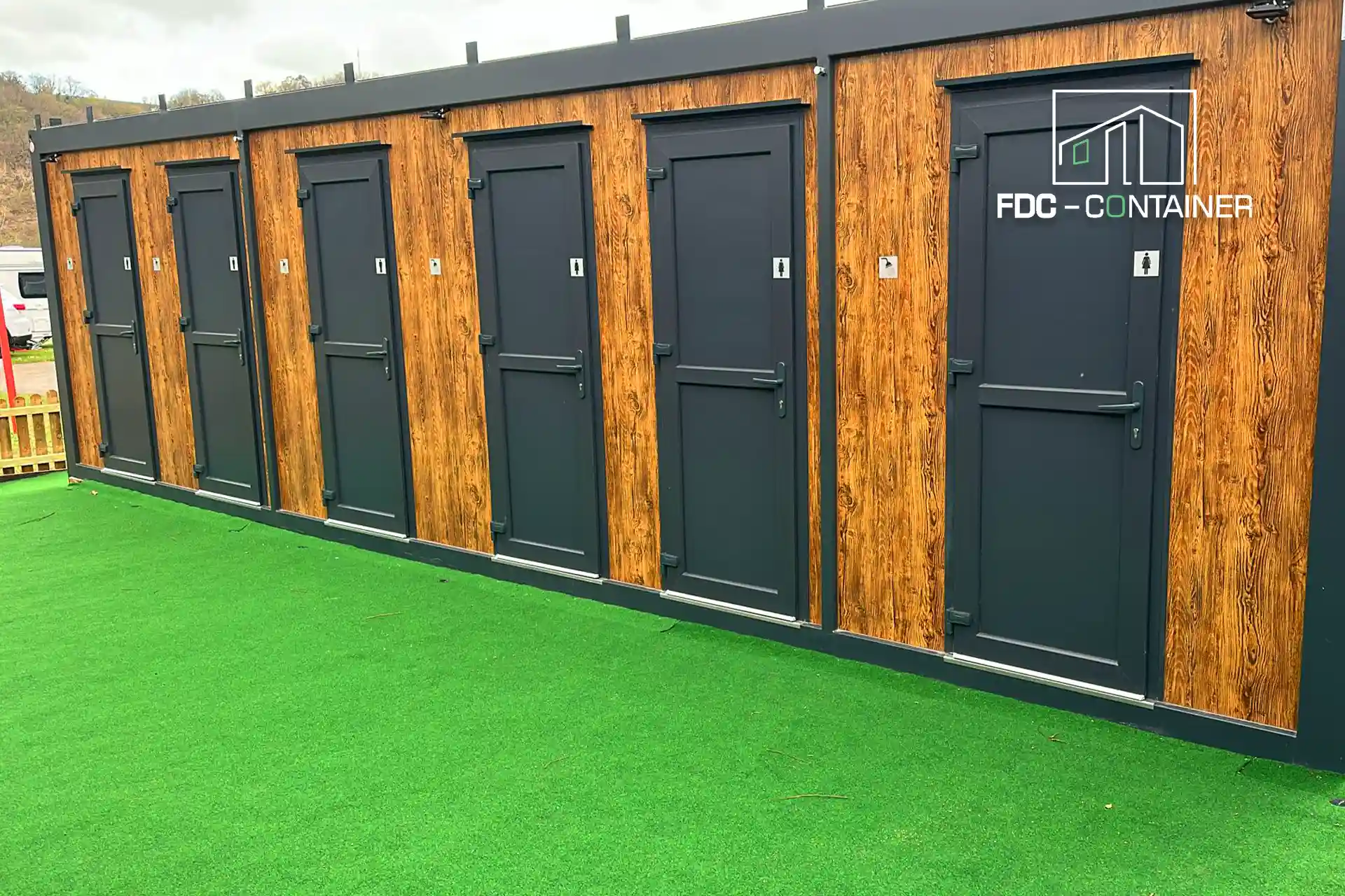 Sanitärcontainer mit Kabinentüren in Anthrazit und Holzoptik auf Kunstrasen – FDC Container Sanitärcontainer mit Kabinentüren in Anthrazit und Holzoptik auf Kunstrasen – FDC Container